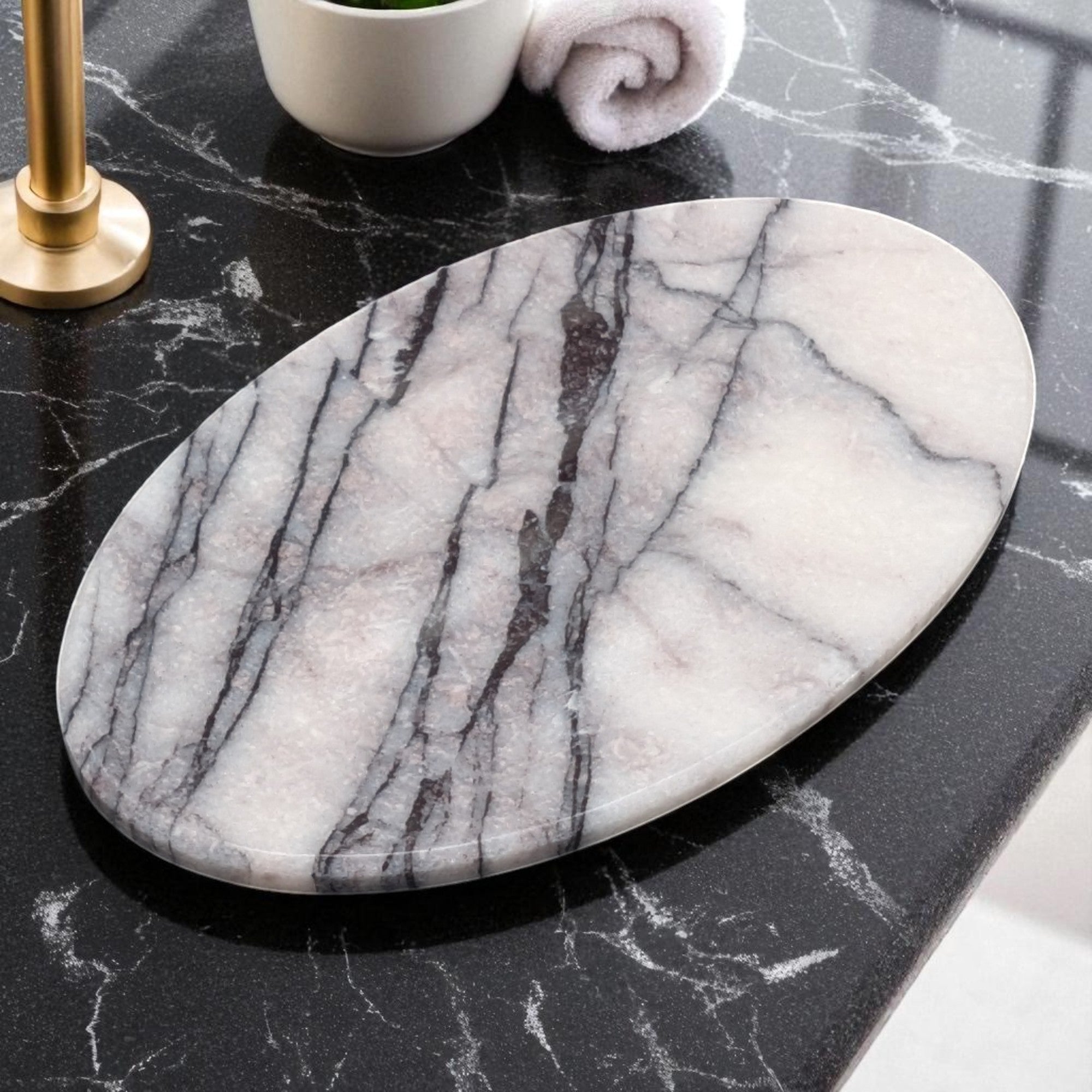 Handcrafted oval white marble soap dish with dramatic grey veining placed on a black marble bathroom countertop beside a rolled white towel and gold fixture