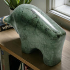 Green stone sculpture of a dog on a wooden surface with books and a plant in the background