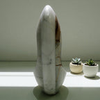 Marble sculpture on a reflective surface with two small potted plants in the background.