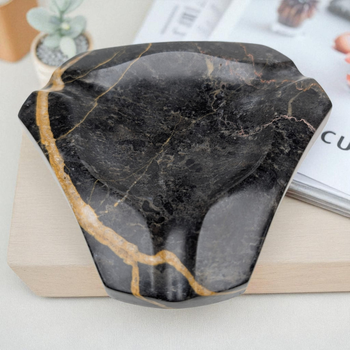 Geometric marble ashtray in black stone with gold veining, resting on a book beside a small succulent